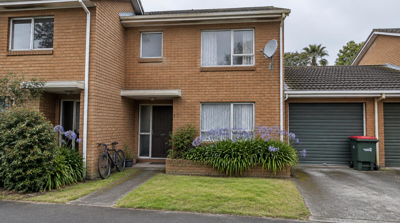 Small 1980s two-storey brick townhouse unit in Remuera with aluminium windows, net curtains, agapanthus in a low brick planter, a wheelie bin and a satellite dish under an overcast sky